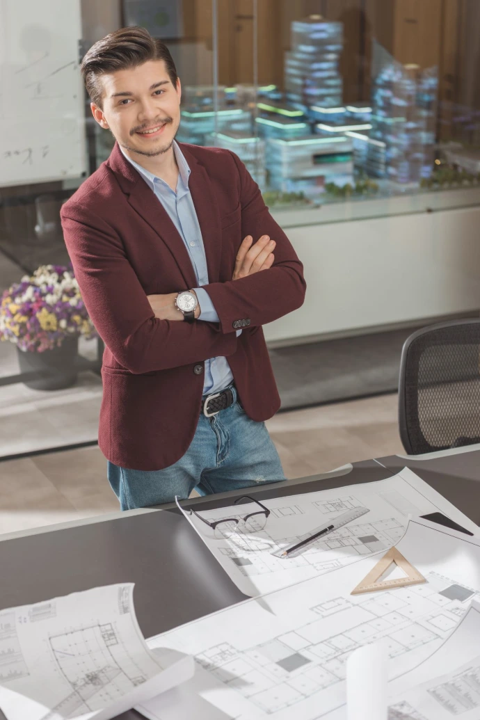 smiling young architect with crossed arms near workplace at office