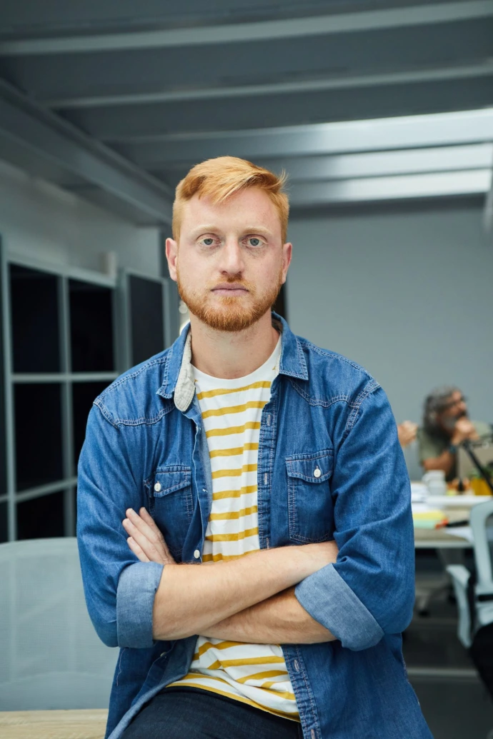 Vertical young red-haired programmer or businessman in modern office looking seriously at camera.